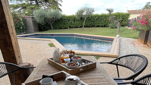 A wooden table with trays of breakfast items, including coffee cups and a teapot, set against a backdrop of a swimming pool and lush greenery.