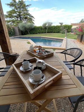 Una mesa de madera con bandejas de alimentos para el desayuno, incluyendo tazas de café y una tetera, con vista a una piscina y abundante vegetación.