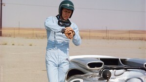 A person in a light blue racing suit standing next to a vintage race car with a number 7 on its side.