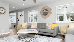 Un salon moderne avec un canapé gris, des coussins jaunes à motifs, une table basse blanche et un tapis en marbre. On aperçoit également, à l’arrière-plan, une grande horloge murale, un miroir en forme de soleil et un coin repas.
