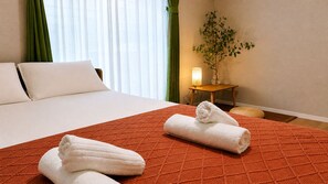 Habitación de hotel con cama, colcha roja con textura, dos toallas enrolladas, mesita de noche con lámpara y planta en maceta.