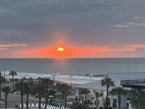 A beachside view with a sunset, ocean waves, and a building with palm trees.