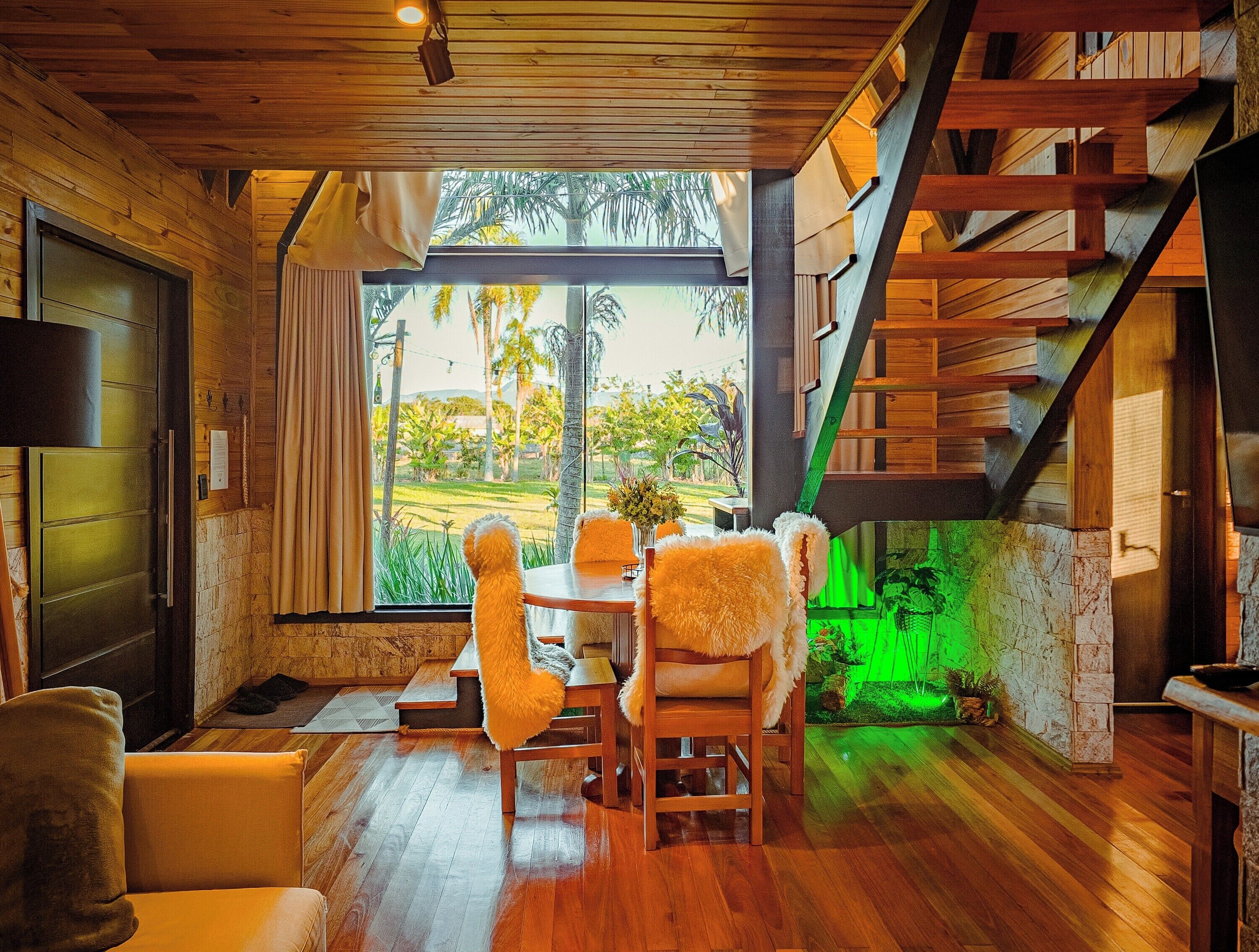 Quarto com piso de madeira, mesa de jantar com cadeiras, sofá, escada e vista para o verde lá fora.