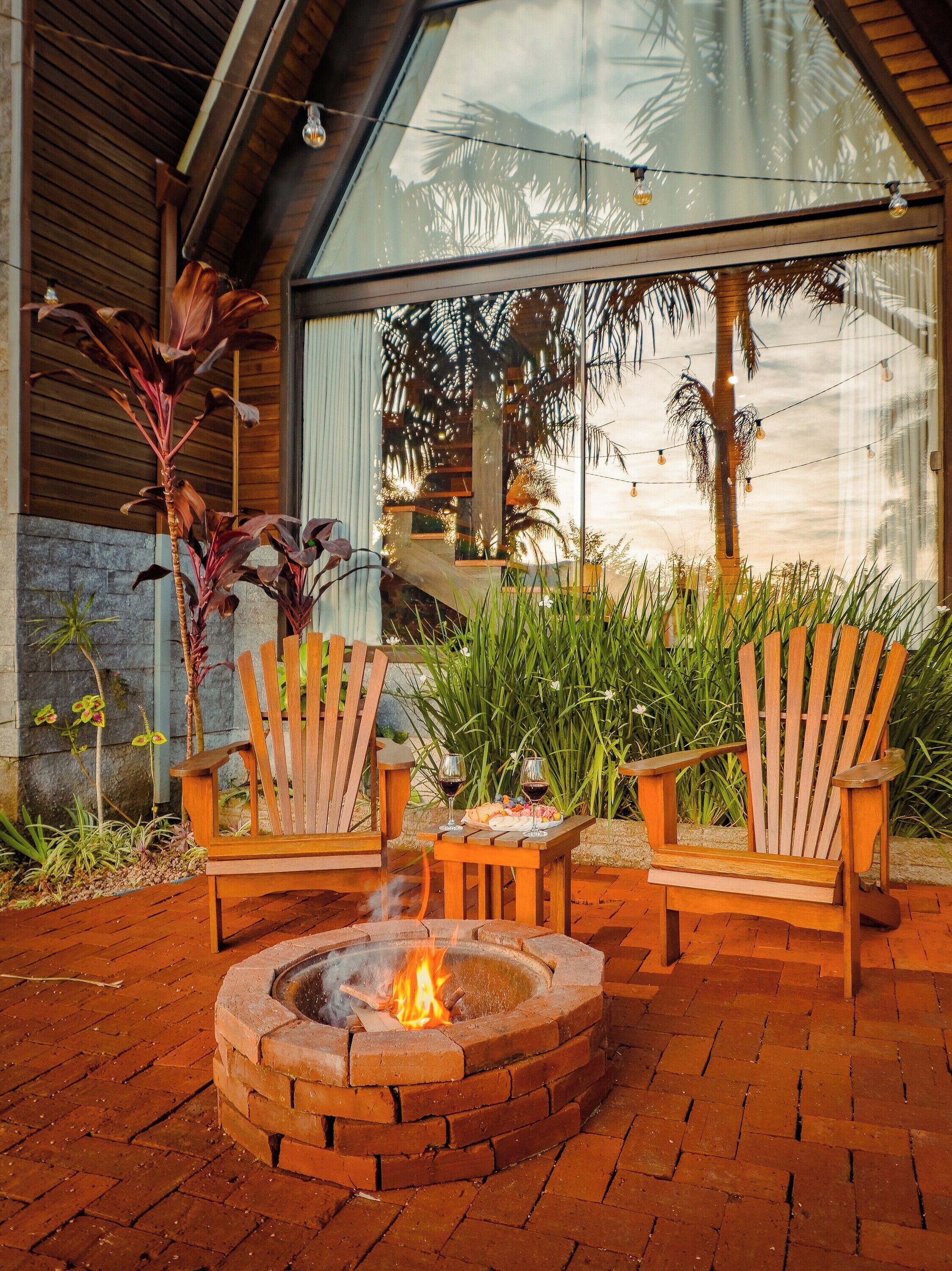 A fire pit with two wooden chairs and a table, surrounded by plants and a brick floor.