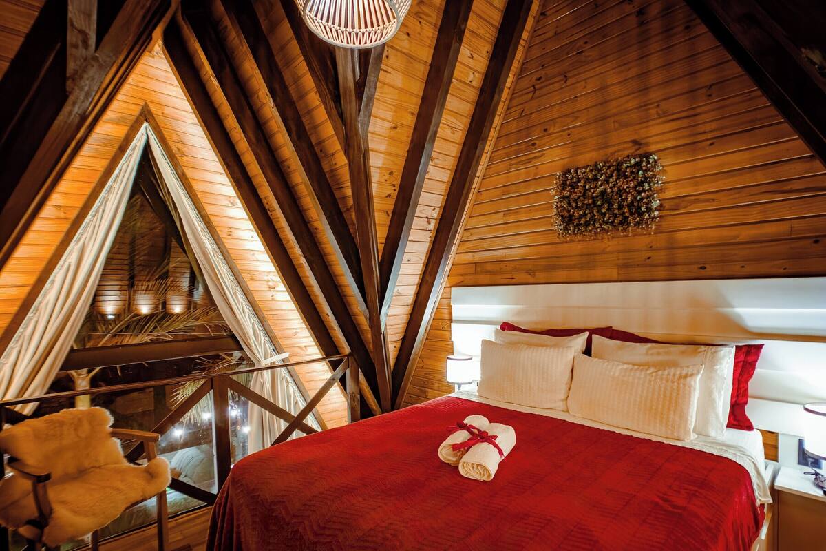 Quarto aconchegante com teto de madeira, cama com roupa de cama vermelha e uma cadeira pequena.