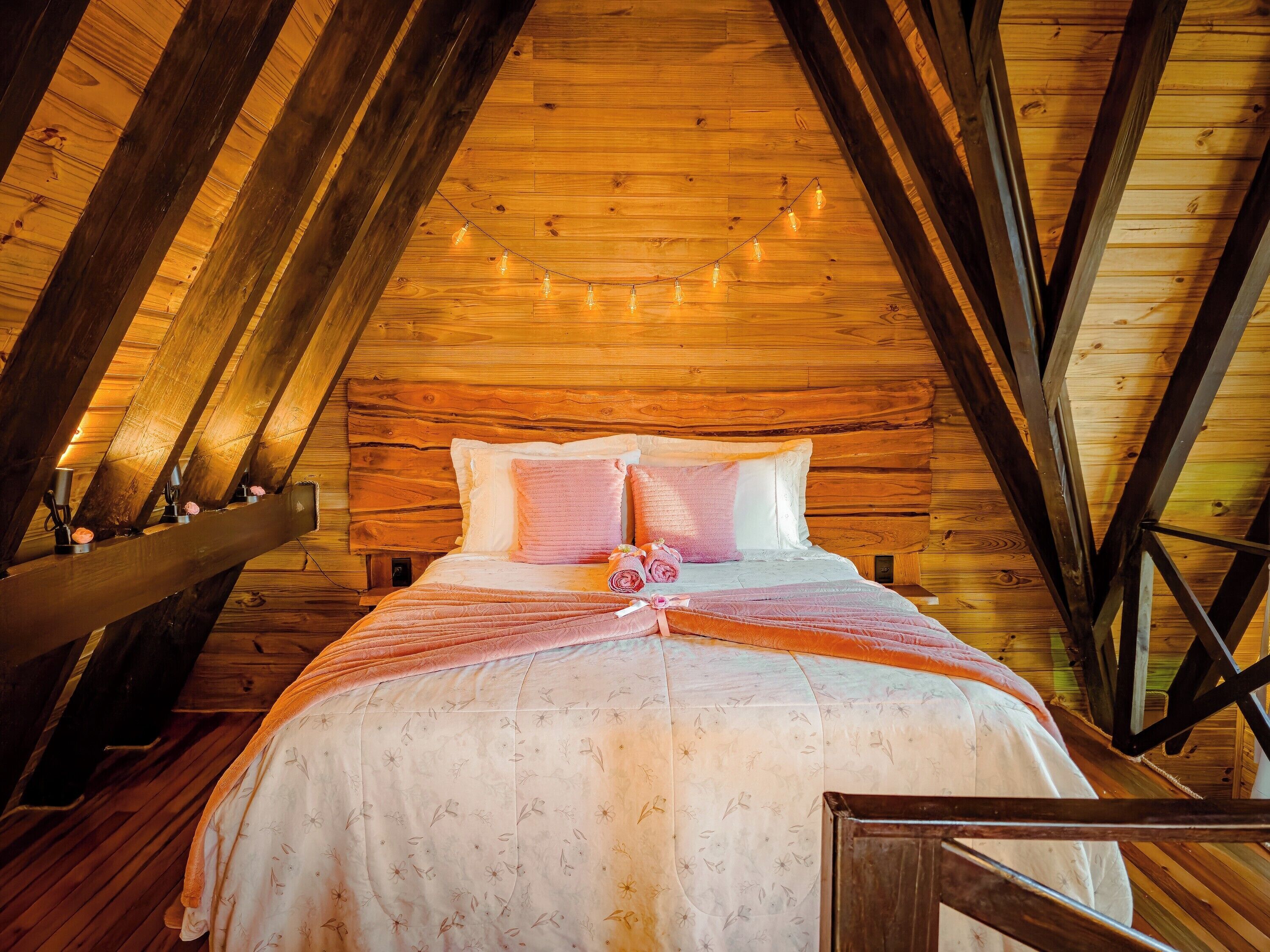 Un dormitorio acogedor en el ático, con techo inclinado de madera, ropa de cama con estampado floral y luces colgantes.