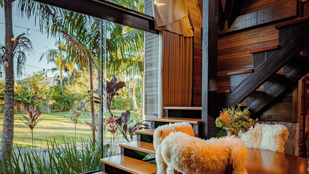 A cozy interior with a wooden staircase, a dining table with a fluffy chair, and a view of a lush garden. - AriMara Chalés & Ecoturismo (Mampituba)