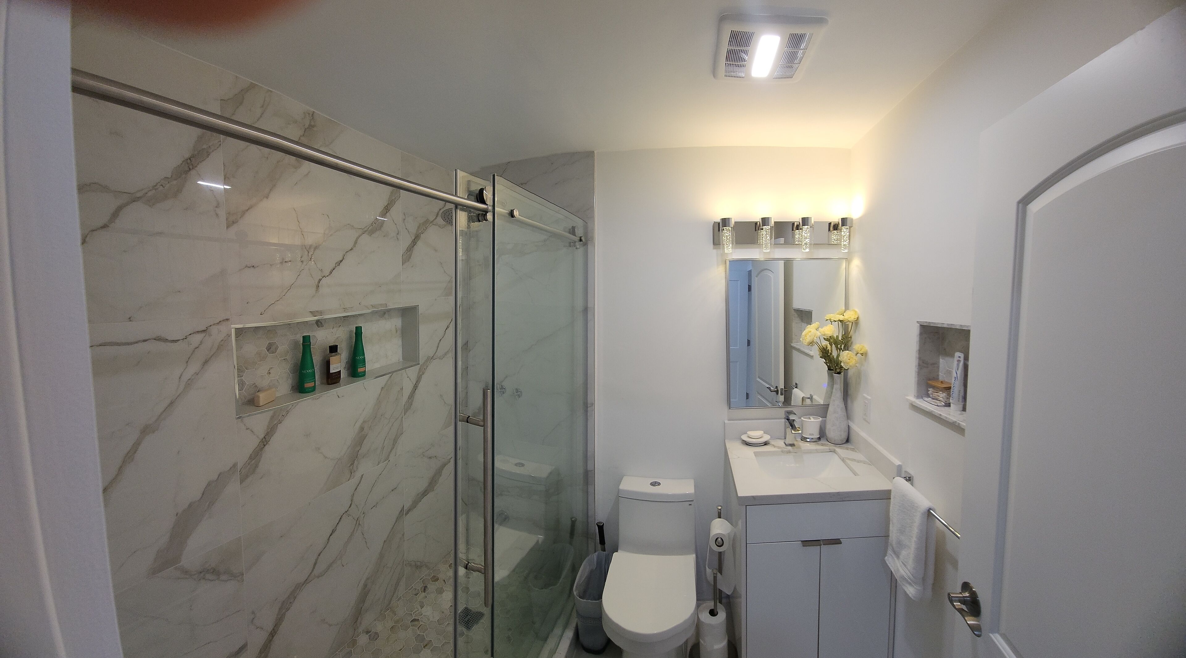 A modern bathroom with a glass-enclosed shower, marble walls, a toilet, and a vanity with a mirror and shelf.