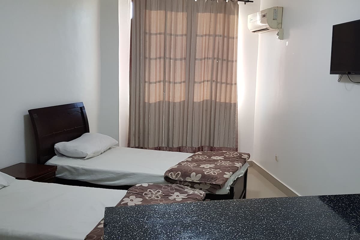 A hotel room with two beds, a flat-screen TV mounted on the wall, an air conditioning unit, and a window with curtains.