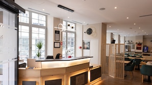 A hotel reception area with a wooden reception desk, a clock on the wall, and a seating area with tables and chairs. - HOTEL SAINT AUGUSTIN (Strasbourg)