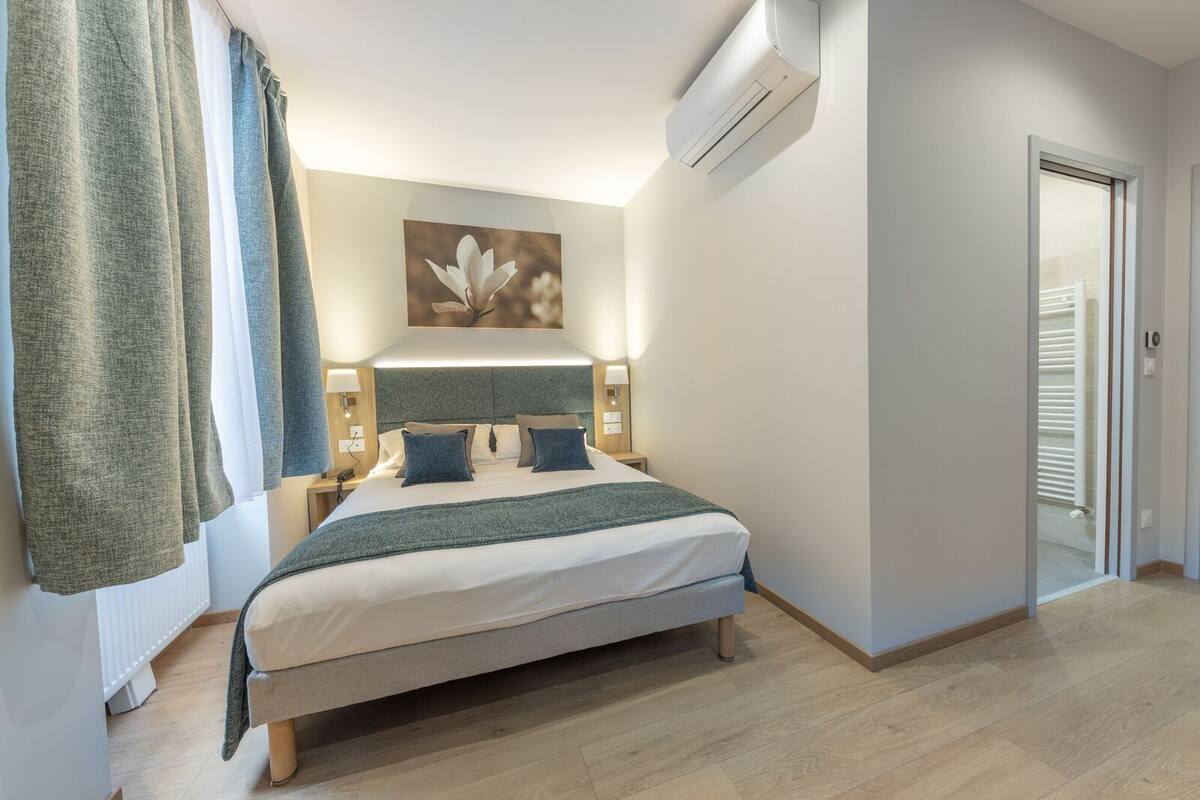 Quarto bem arrumado com cama, abajures, ar condicionado e uma porta que dá para outro cômodo.