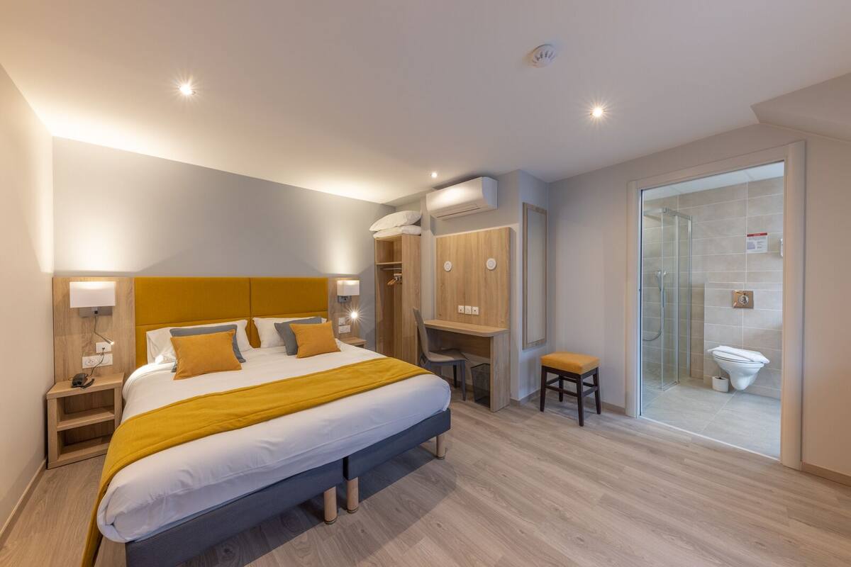 Quarto de hotel moderno com uma cama grande, uma escrivaninha, chuveiro e vaso sanitário.