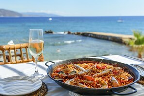 Pöydällä on mereneläinpaella ja lasi valkoviiniä, ja pöydästä avautuu näkymä rannikolle.