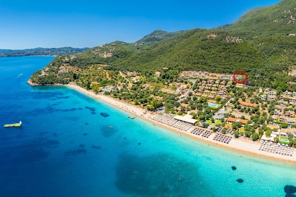 Vista aérea de un pueblo costero con playa, aguas azules cristalinas y colinas verdes exuberantes al fondo.