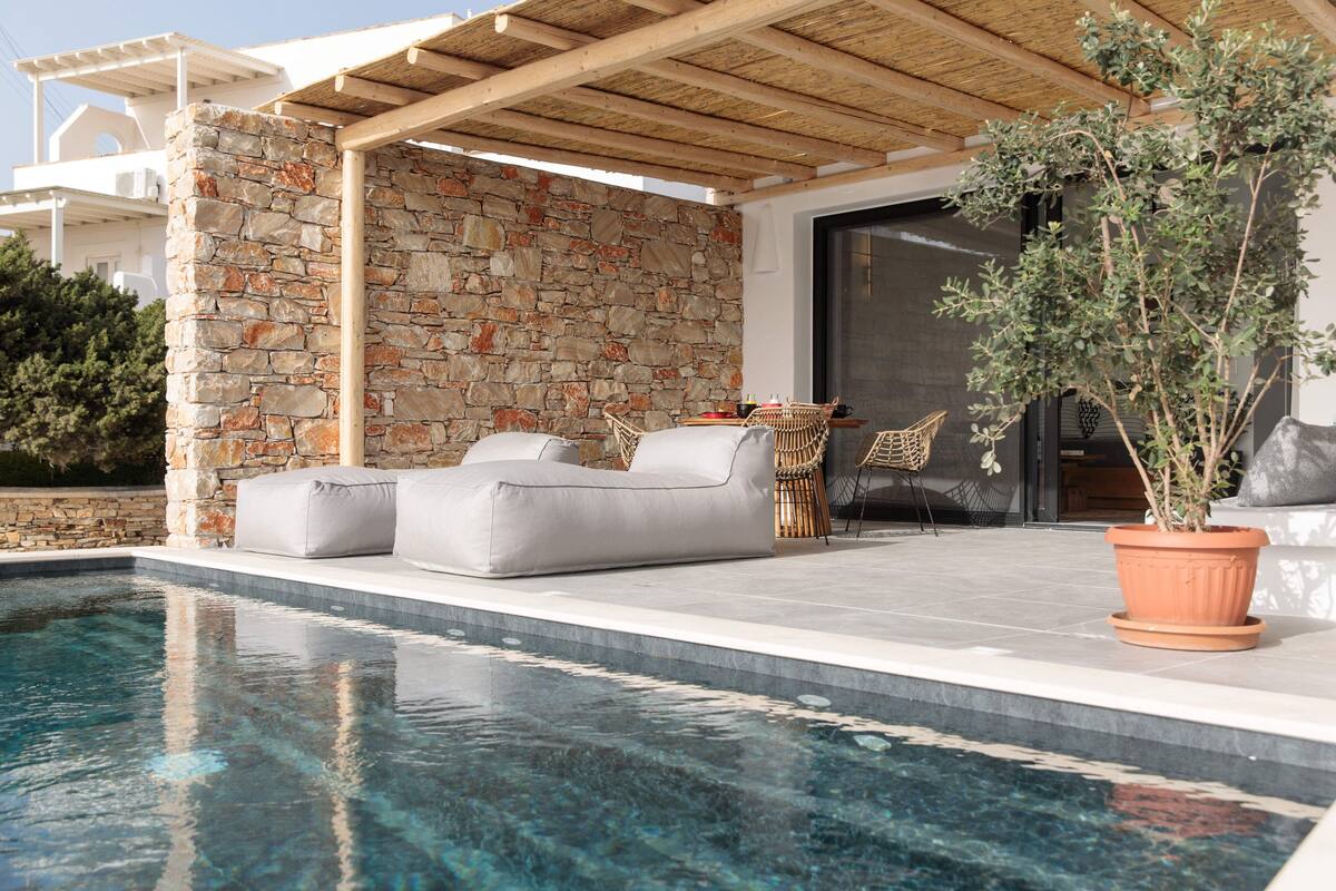 A poolside area with a stone wall, a wooden pergola, and a seating area with cushions and a table.