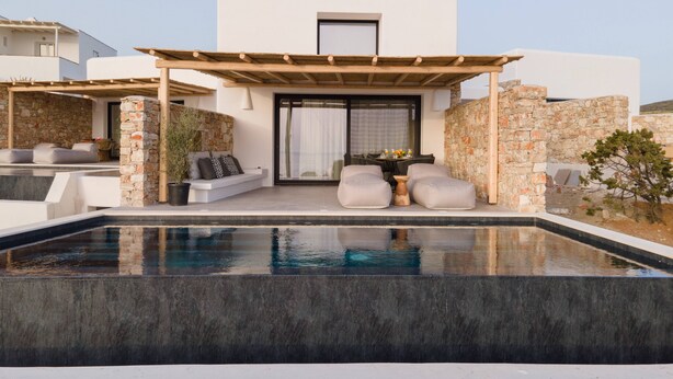 Un área de piscina moderna al aire libre con un muro de piedra, una pérgola de madera y una zona de estar con cojines y una mesa.