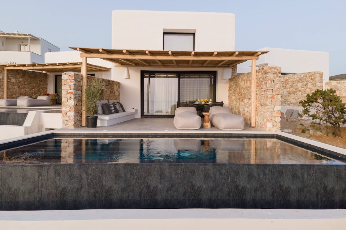 A modern outdoor pool area with a stone wall, a wooden pergola, and a seating area with cushions and a table.