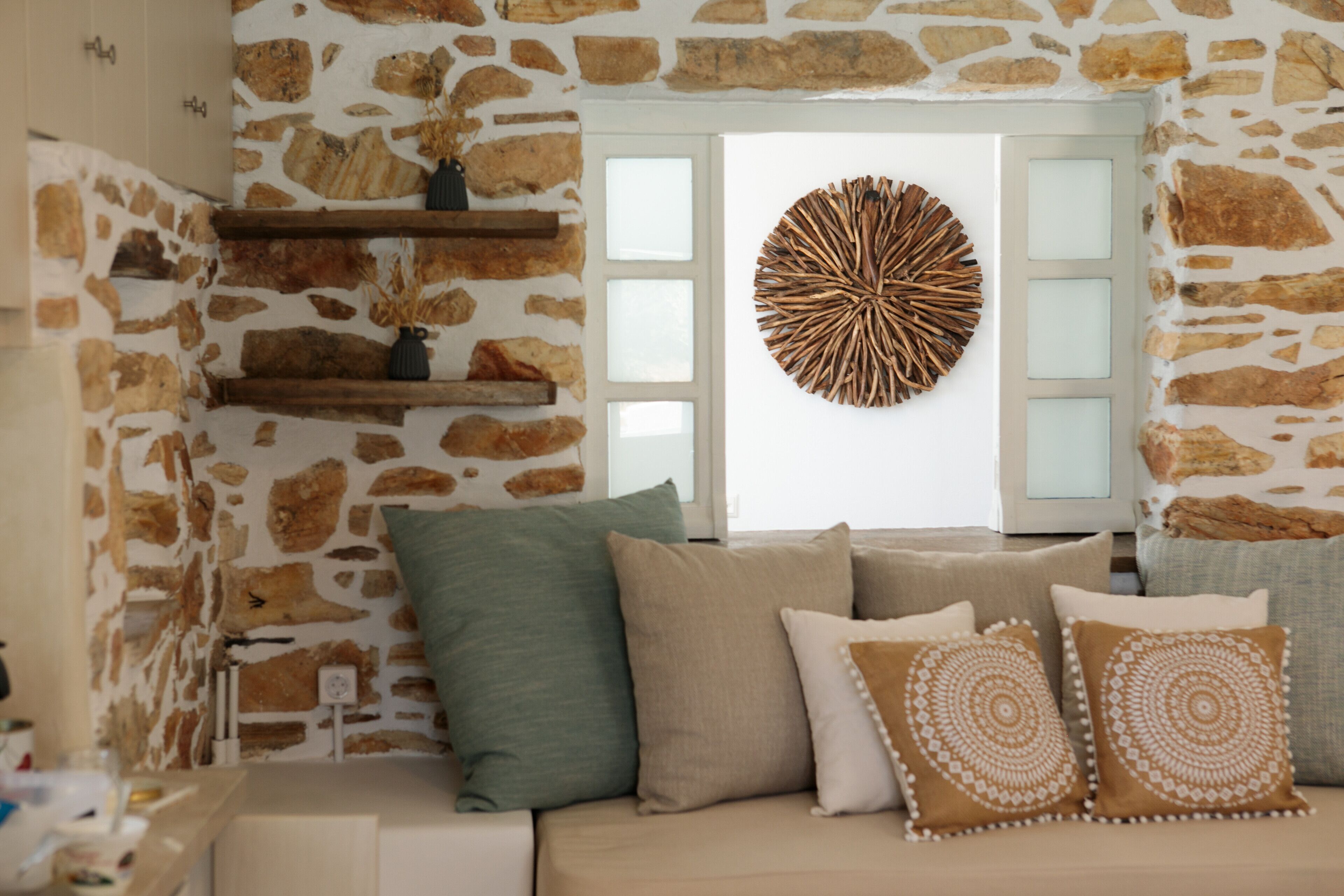A room with a stone wall, a wooden shelf with decorative items, a round wall art piece, and a beige sofa with patterned cushions.