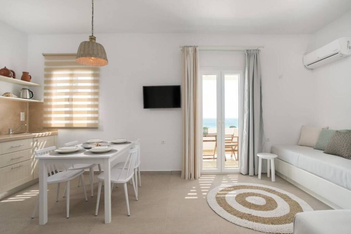 A modern, well-lit room with a dining area, a flat-screen TV, and a view of the sea through a sliding glass door.