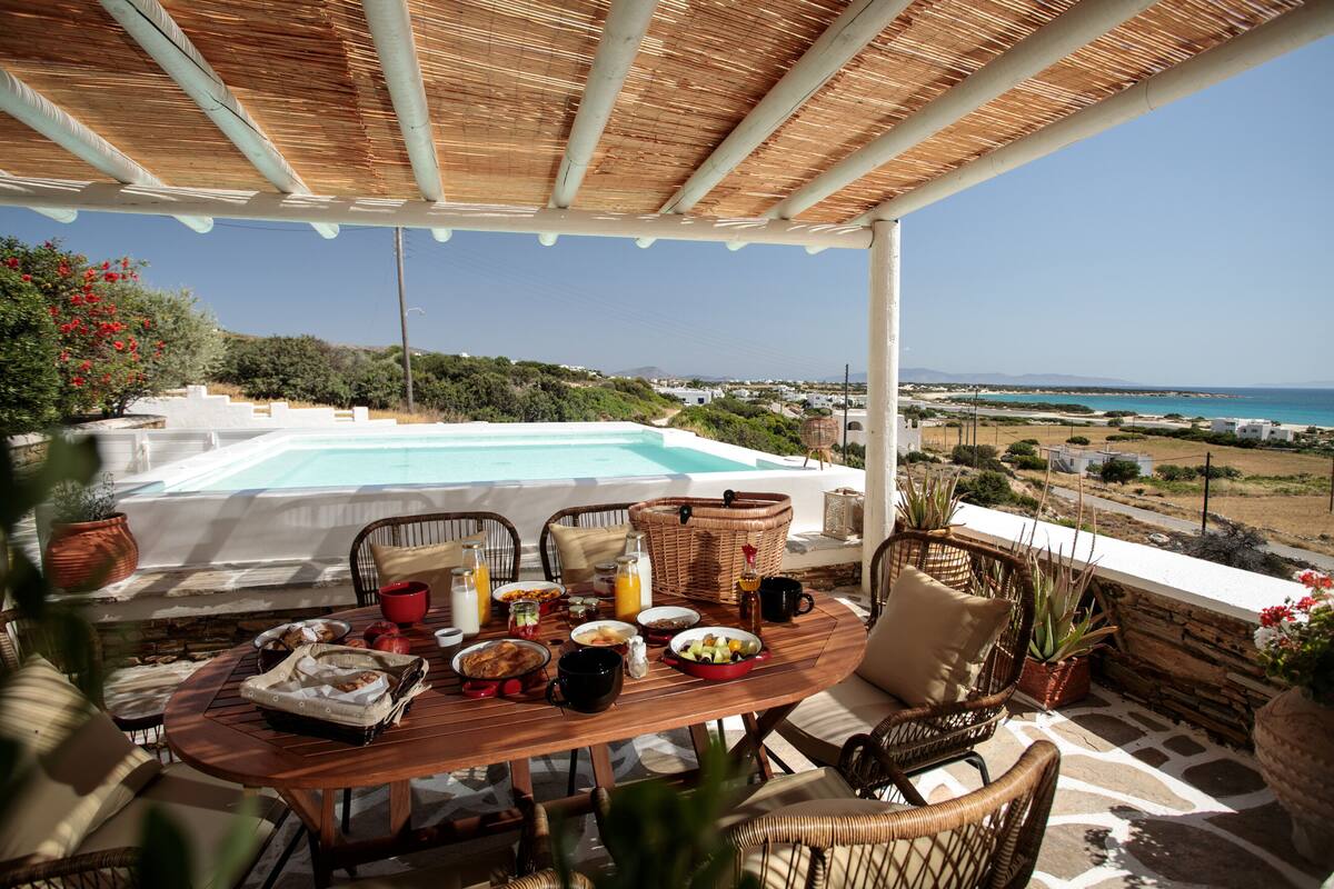 A dining area with a view of a pool and the sea, featuring a table set with breakfast items and wicker furniture.