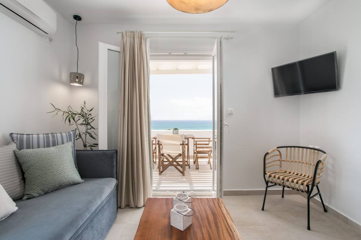 A modern living room with a sofa, a wooden coffee table, a rattan chair, a television mounted on the wall, and a view of the beach through an open door.