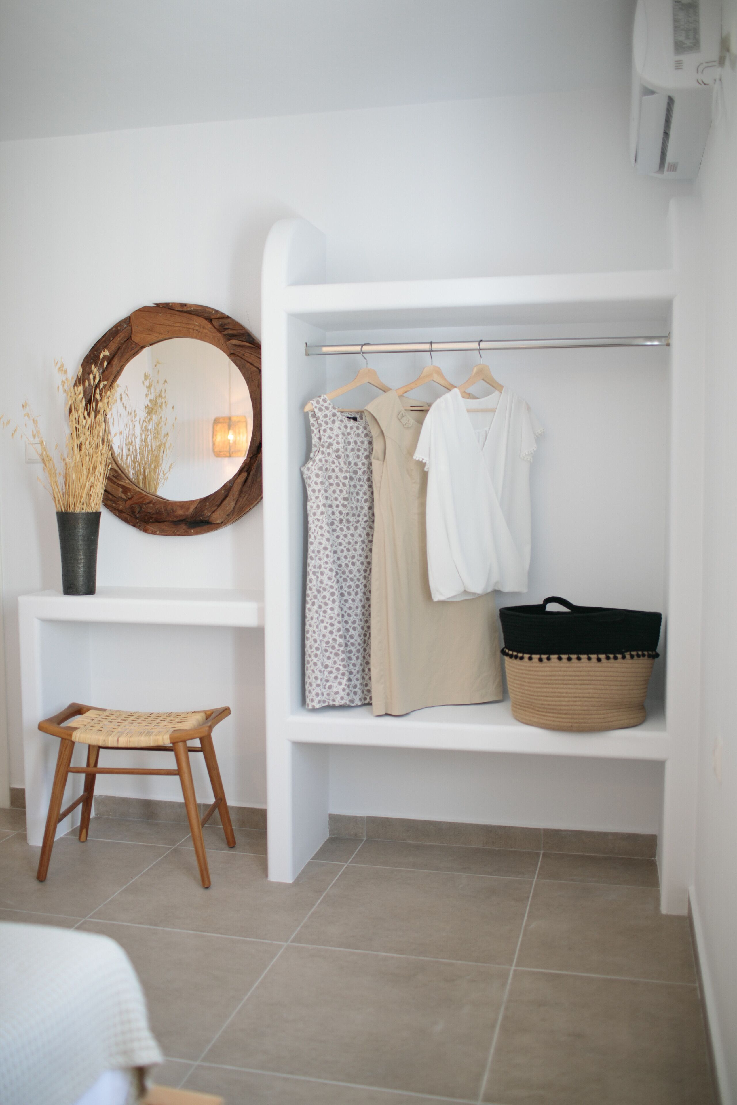 Una habitación blanca con un espejo de marco de madera, una estantería con ropa, una cesta y un taburete pequeño.