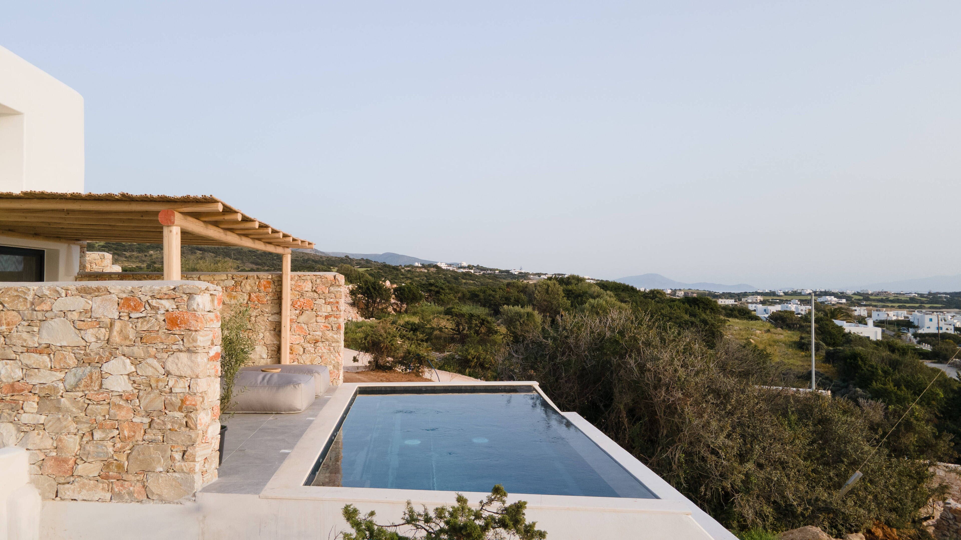A stone-walled infinity pool with a pergola, overlooking a lush landscape and distant buildings.