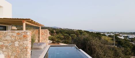A stone-walled infinity pool with a pergola, overlooking a lush landscape and distant buildings.