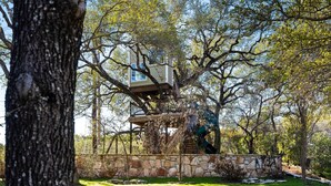 Une cabane perchée dans un arbre, dotée d’un mur en pierre et d’une terrasse en bois, entourée d’arbres et de verdure.