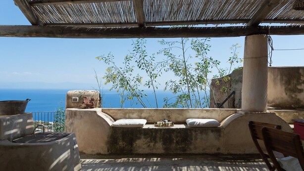 A concrete outdoor seating area with a view of the sea, a metal grill, and a wooden chair. - Queen's Hawk Case (Filicudi Porto)
