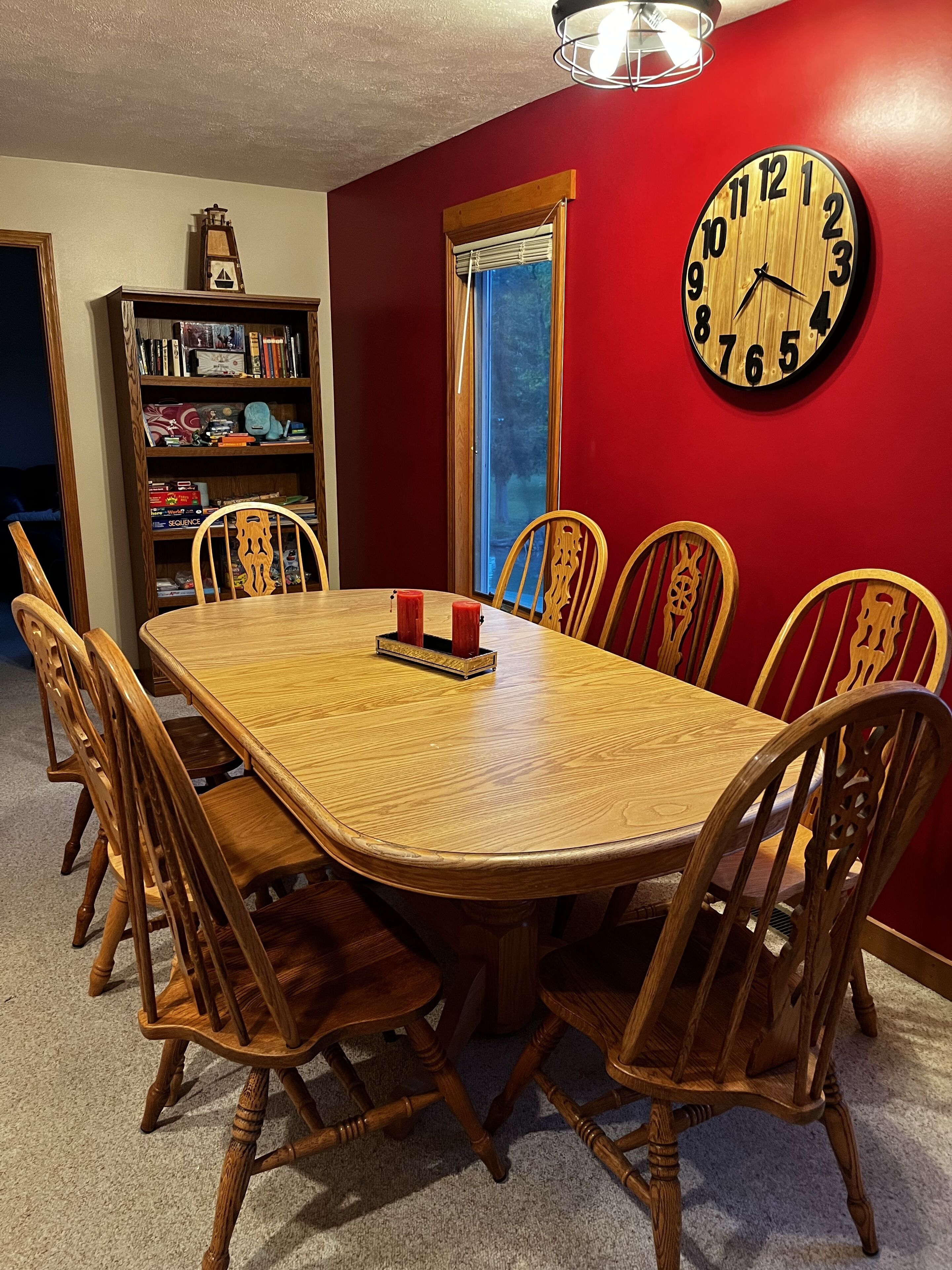 Comedor con mesa y sillas de madera, una estantería, un reloj y una ventana con cortinas.