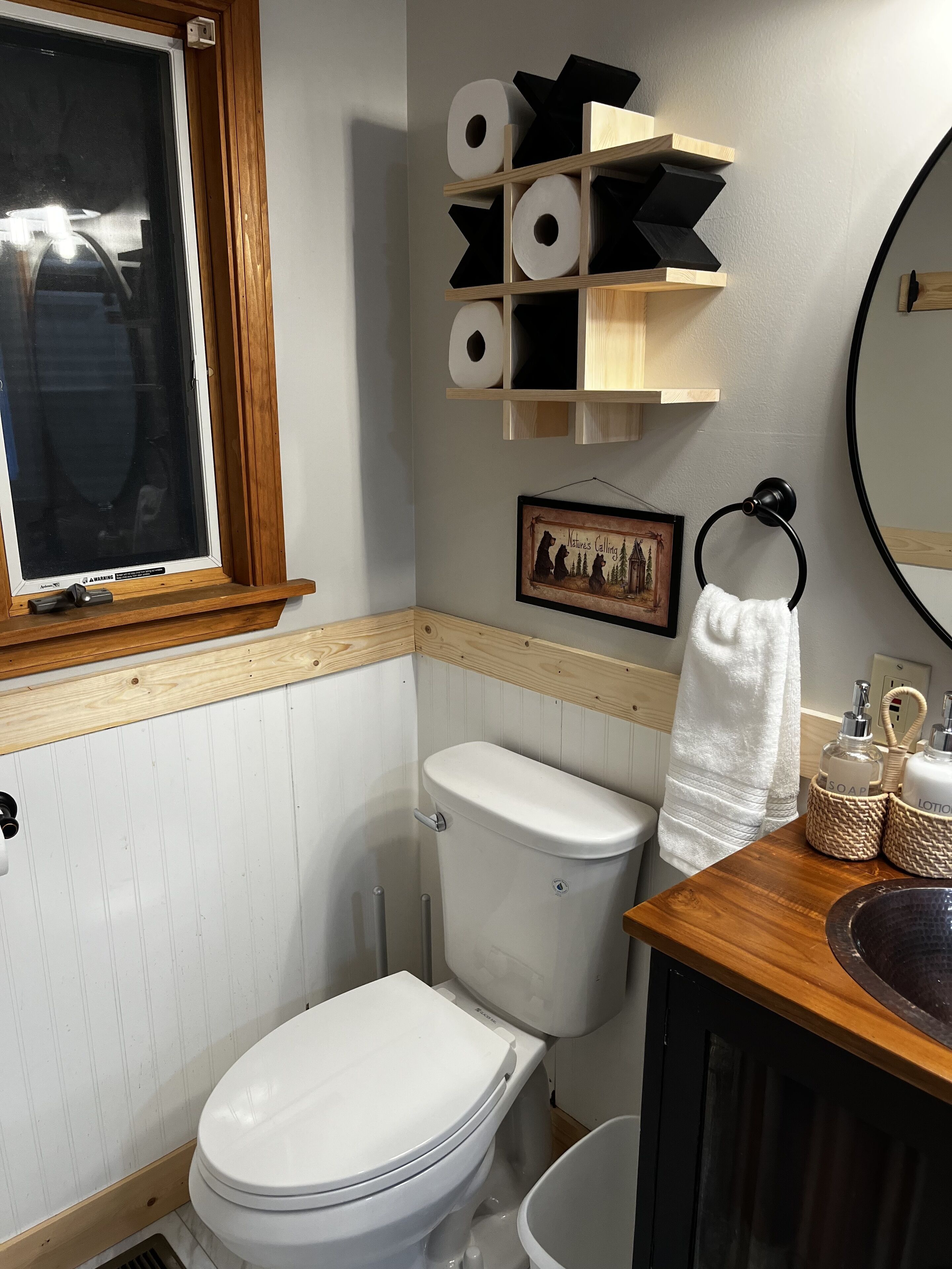 A bathroom with a toilet, a wooden shelf with toilet paper, a framed picture, and a towel rack.