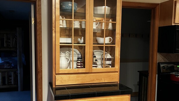 Una cocina con armarios de madera, encimera negra y un aparador con vidrio que muestra vajilla.