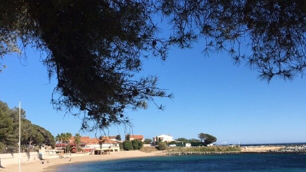 A coastal beach with clear blue water, a sandy shore, and buildings in the background. - Studio Proche Mer, son Jardinet et sa Place de Parking Privés (Roquebrune-sur-Argens)