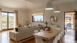 A modern living room with a sofa, dining table, and chairs. There is a view of the outdoors through large windows. - Canyon 2 villa with private pool (Rethymno)