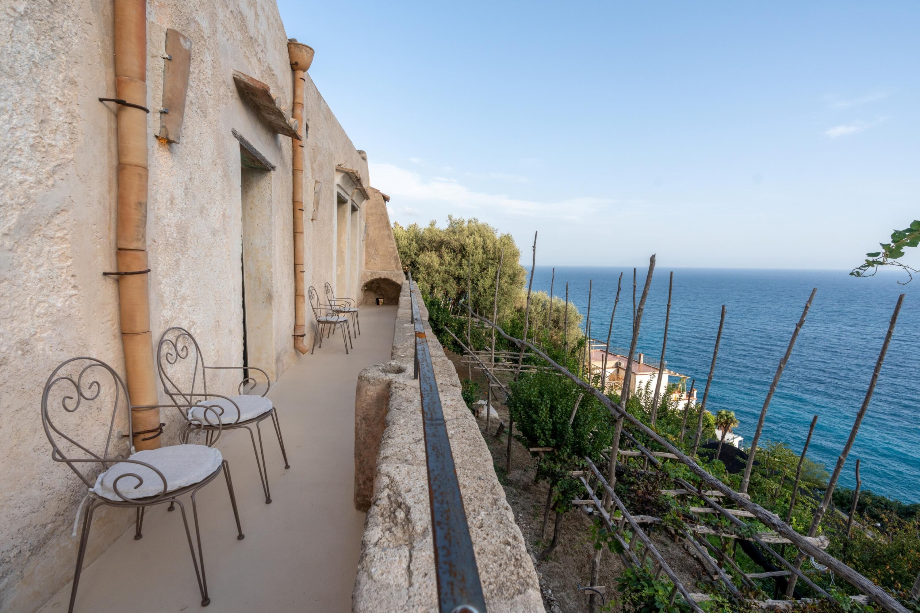 Une terrasse en bord de mer, avec des murs en pierre, des chaises en métal et une vue sur l’océan.