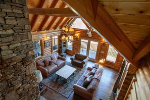 Cozy cabin interior with a high ceiling, exposed beams, and a stone wall. Features a seating area with a sofa, armchairs, and a fireplace.
