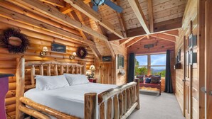 A log cabin bedroom with a wooden bed, a nightstand, a window with a view, and a wooden ceiling.