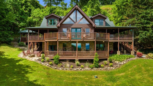 A two-story log cabin with a balcony on each floor, surrounded by a well-maintained lawn and dense forest.