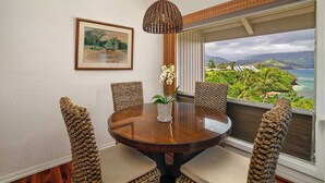 Uma mesa de madeira redonda com um vaso branco e orquídeas, rodeada por cadeiras de fibra, num quarto com uma janela ampla com vista para uma paisagem tropical.