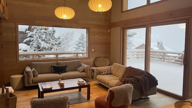 Ett mysigt vardagsrum med en soffa, fåtölj och soffbord, stora fönster med utsikt över ett snötäckt landskap, träväggar och hängande taklampor.