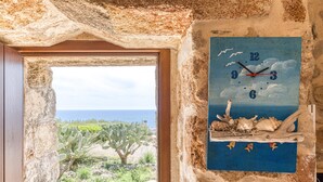 A rustic room with a view of the sea through a doorway, a wall clock with a beach scene, and a cactus plant.