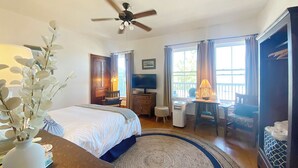 Ein Hotelzimmer mit Bett, Schreibtisch, Sessel, Fernseher, Deckenventilator und einem Fenster mit Vorhängen.