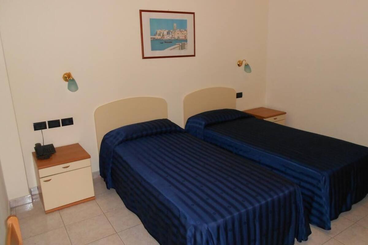 A room with two beds, a nightstand with a lamp, a small table, and a picture on the wall.