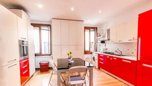 A compact kitchen with white and red cabinets, a dining table with chairs, and a window providing natural light. - Ca’ Lunetta  (Venezia)