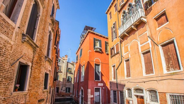 A narrow street with buildings on either side, featuring a mix of brick and painted facades, and a clear blue sky above. - Ca’ Lunetta  (Venezia)