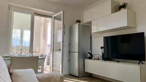 A modern living room with a flat-screen TV, a built-in kitchen area, and a sliding glass door leading to a balcony.