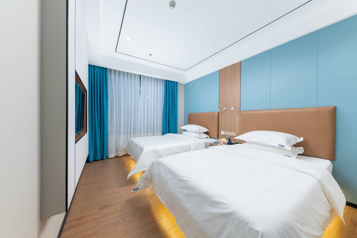 A hotel room with two beds, a wooden floor, a blue accent wall, and a large window with curtains.