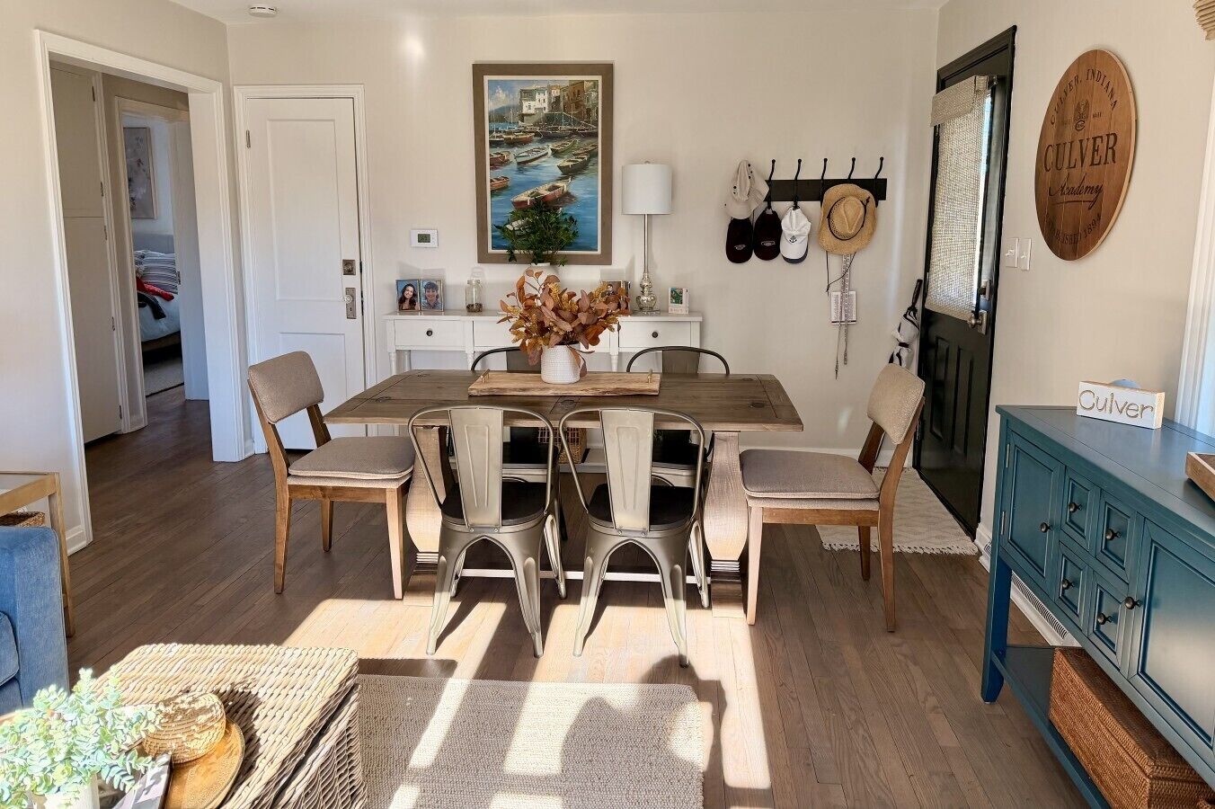 Een eetgedeelte met een houten tafel, zes stoelen, een vaas met gedroogde bloemen, een schilderij boven de tafel en een hoedenrek aan de muur.