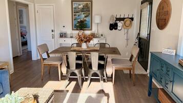 Een eetgedeelte met een houten tafel, zes stoelen, een vaas met gedroogde bloemen, een schilderij boven de tafel en een hoedenrek aan de muur.